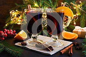 Two glasses of mulled wine, cinnamon and oranges on a festive Christmas background with branches of a pine tree with a garland.