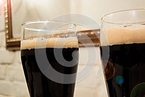 Two glasses poured with dark beer or soft drink with foam head. Close up image against white brick wall in bar or pub