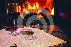 Two glasses of red wine against the background of a burning fireplace in hard reflective lighting. Romantic relaxed dinner by the