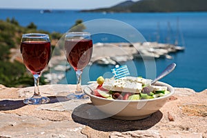 Two glasses of red wine and bowl of greek salad with greek flag on by the sea view, summer greek holidays concept.