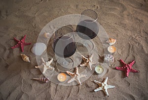 Two glasses of red wine, burning candles, seashells and starfishes on sand beach.