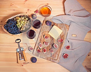 Two glasses of red wine, corkscrew, bunches of grape in wicker basket, figs, cheese and  honey in glass bowl on wooden table