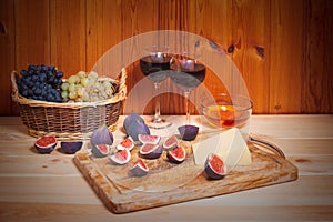 Two glasses of red wine, figs, cheese, bunches of grape and bowl of honey on wooden table.