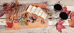 Two glasses of red wine, figs, cheese, grape, bread, black olives and autumn leaves on aged wooden table.