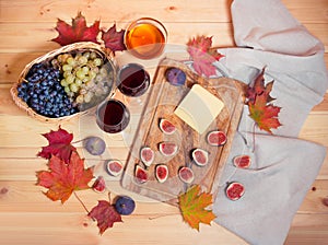 Two glasses of red wine, figs, piece of cheese, bunches of grape, honey and autumn maple leaves on wooden table