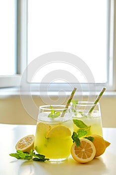 Two glasses of refreshing iced lemonade with lemon slices, mint leaves and ice on kitchen counter. Non alcoholic mojito drink on