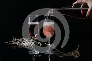 Two glasses of rose champagne and Christmas or New Year decoration on black background. Woman`s hand holds a bottle and pouring