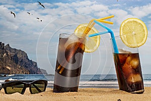 Two glasses of soda with ice and straws on the beach and and sunglasses with blue sky and seagulls flying in Tenerife, Canary