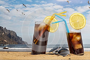 Two glasses of soda with ice and straws on the beach and sunglasses with blue sky and seagulls flying in Tenerife, Canary Islands