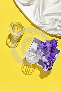 Two glasses with water on yellow background with white silk cloth and wooden tray with violet iris flowers. Summer refreshment