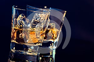 Two glasses of whiskey with ice cubes on black background with light tint blue