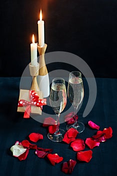 Two glasses with white champagne, gift box and petals of red roses on the background of candles