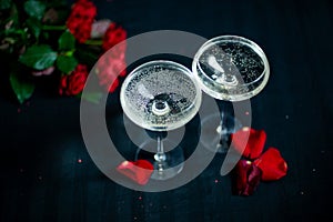 Two glasses with white champagne and petals of red roses on the black background