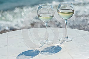 Two glasses of white wine on a white table on the beach near the water. Sparkling sun glare on the water,