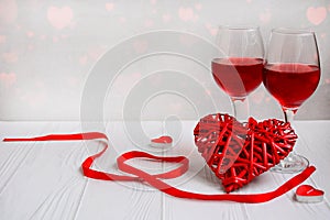 Two glasses of wine and a wicker heart on a light background with hearts. Valentine`s Day