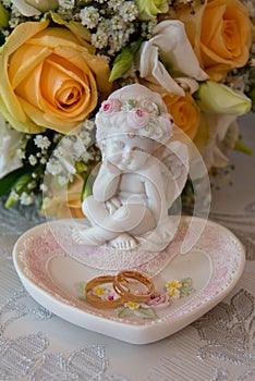 Two gold wedding rings lie on a platter in a rose shape with the angel sculpture near the bride's bouquet