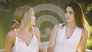 Two good friends are having a heart to heart conversation out on a park bench. Girls wearing white dresses discuss