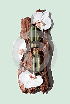Two green glass cosmetic bottles on textured tree bark podium with white orchid flowers on pastel mint background.