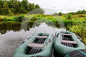 Two green rubber boats are lying on the green grass on the shore of a lake overgrown with grass.