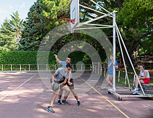 Two guys playing basketball on an outdoor basketball court and two girls watching