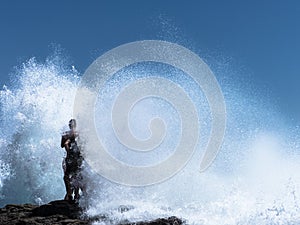 Two guys standing on a rock in the waves.