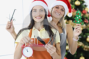 Two hairdresser women in santa claus hats hold scissors and combs with alarm clock on background of New Year tree