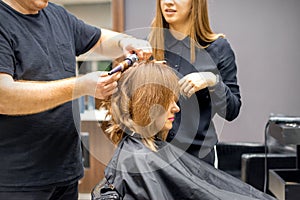 Two hairstylists make curls hairstyle of long brown hair with the curling iron in hairdresser salon.