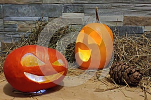 Two halloween jack o lanterns yellow and red on foreground on a hay and brick wall background. with cones on wooden board