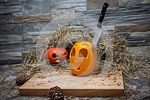 Two halloween pumpkins lie on a hay against the background of a stone wall. The knife is stuck in a yellow pumpkin. Near the cones
