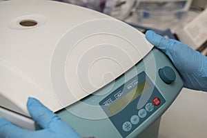 Two hands with blue nitril gloves next to the control panel of a closed centrifuge in a laboratory.