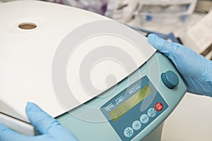 Two hands with blue nitril gloves next to the control panel of a closed centrifuge in a laboratory.