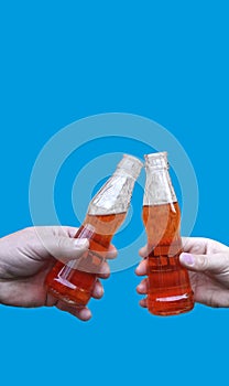 Two hands holding a drink and making toast, clinking bottles with lemonade or beer, isolated on blue background