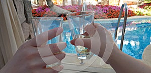 Two hands holding glasses with white vine together over blue pool