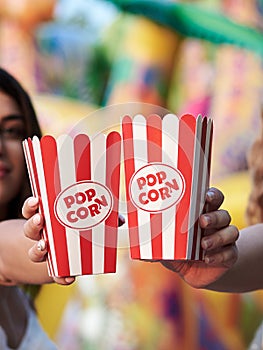 Two hands, holding popcorn in red and white paper boxes, Close-up picture of snack boxes at fun fair in theme park. Summertime