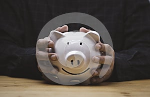 Two hands holding white smile piggy saving bank on wooden table for money save deposit and financial banking concept