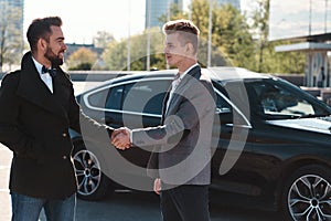 Two handsome businessmen shaking hands in front of the black car standing outdoors