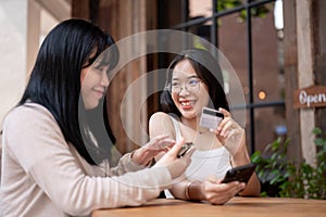 Two happy Asian female friends are talking about the benefits of their credit cards at a cafe