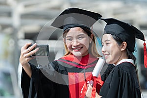 Two happy Asian young beautiful graduate female students with University degree standing and holding diploma taking selfie picture