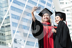 Two happy Asian young beautiful graduate female students with University degree standing and holding diploma taking selfie picture