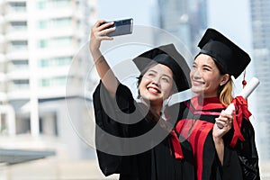 Two happy Asian young beautiful graduate female students with University degree standing and holding diploma taking selfie picture