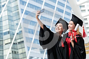 Two happy Asian young beautiful graduate female students with University degree standing and holding diploma taking selfie picture