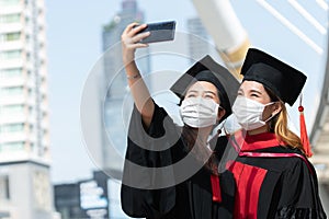 Two happy Asian young beautiful graduate female students wearing face mask standing and taking selfie picture with mobile phone