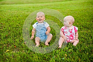 Two happy baby boy and a girl age 9 months old, sitting on the grass and interact, talk, look at each other.