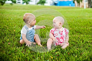 Two happy baby boy and a girl age 9 months old, sitting on the grass and interact, talk, look at each other.