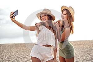Two happy blonde caucasian sisters having fun on the beach during summer, taking selfie with mobile phone, smiling and looking to