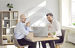 Two happy business men working in office sitting with laptop computer and smiling