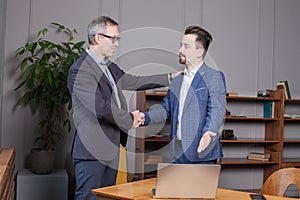 Two happy business partners in blue suits smiling and making handshake in office. Businessman pointing on the laptop