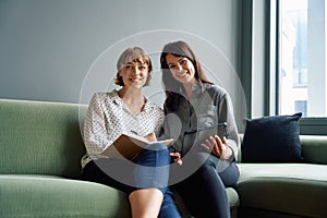 Two happy business women sitting on sofa using digital tablet during meeting in office