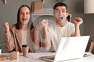 Two happy cheerful Caucasian businesspeople showing clenched fist, looking at camera while sitting in front of laptop screen,