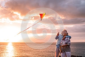 Two happy and cheerful senior having fun enjoying and playing with a flying kite at the beach with the sunset and the sea at the